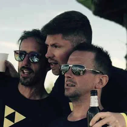 Three men wearing matching Beat Bunker shirts posing outdoors with drinks, smiling and pointing in the distance